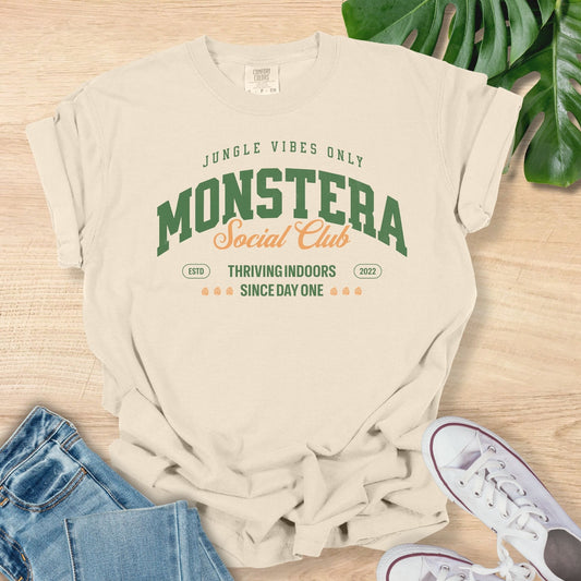 Beige t-shirt with 'Monstera Social Club' text on a wooden surface with jeans and sneakers.