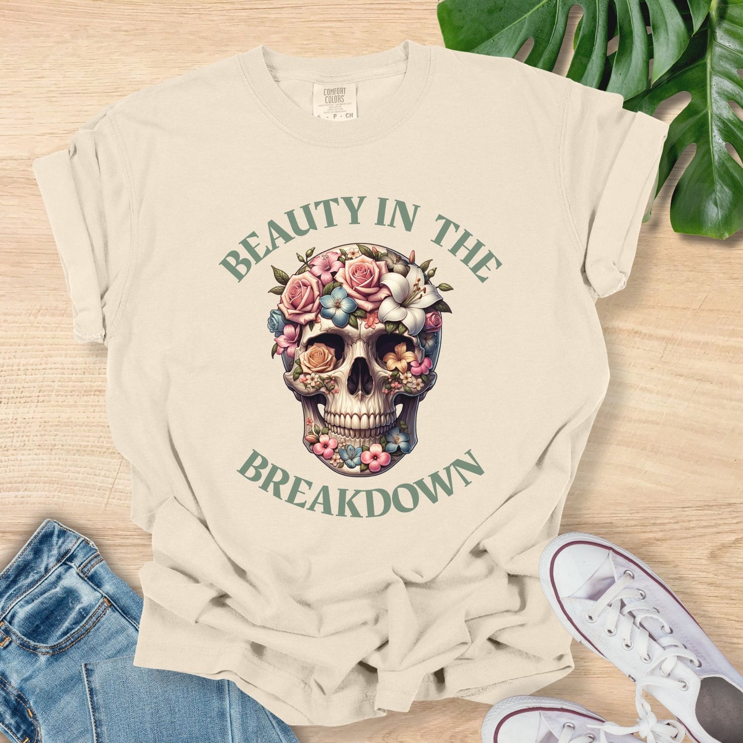 Beige t-shirt with skull and floral design on a wooden surface with jeans and sneakers.