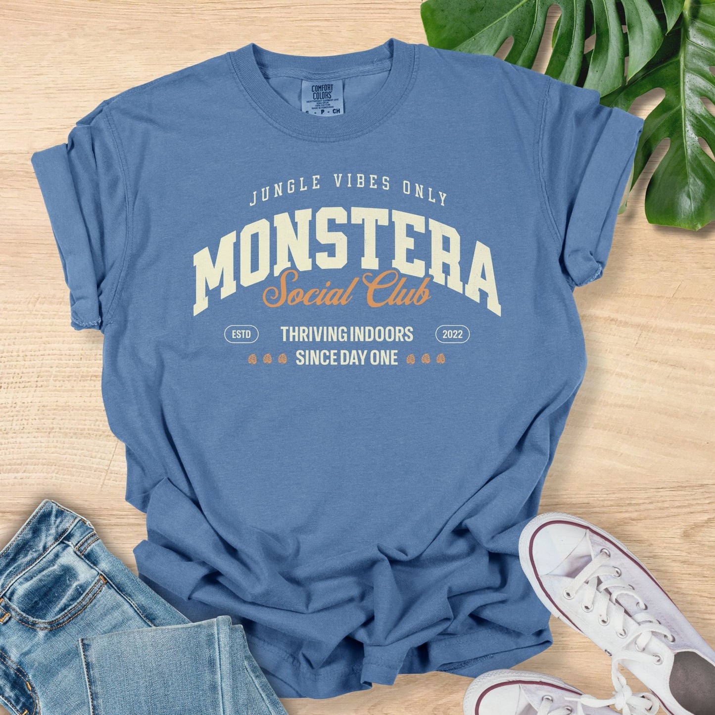 Blue t-shirt with 'Monstera Social Club' text on a wooden surface with jeans and sneakers.