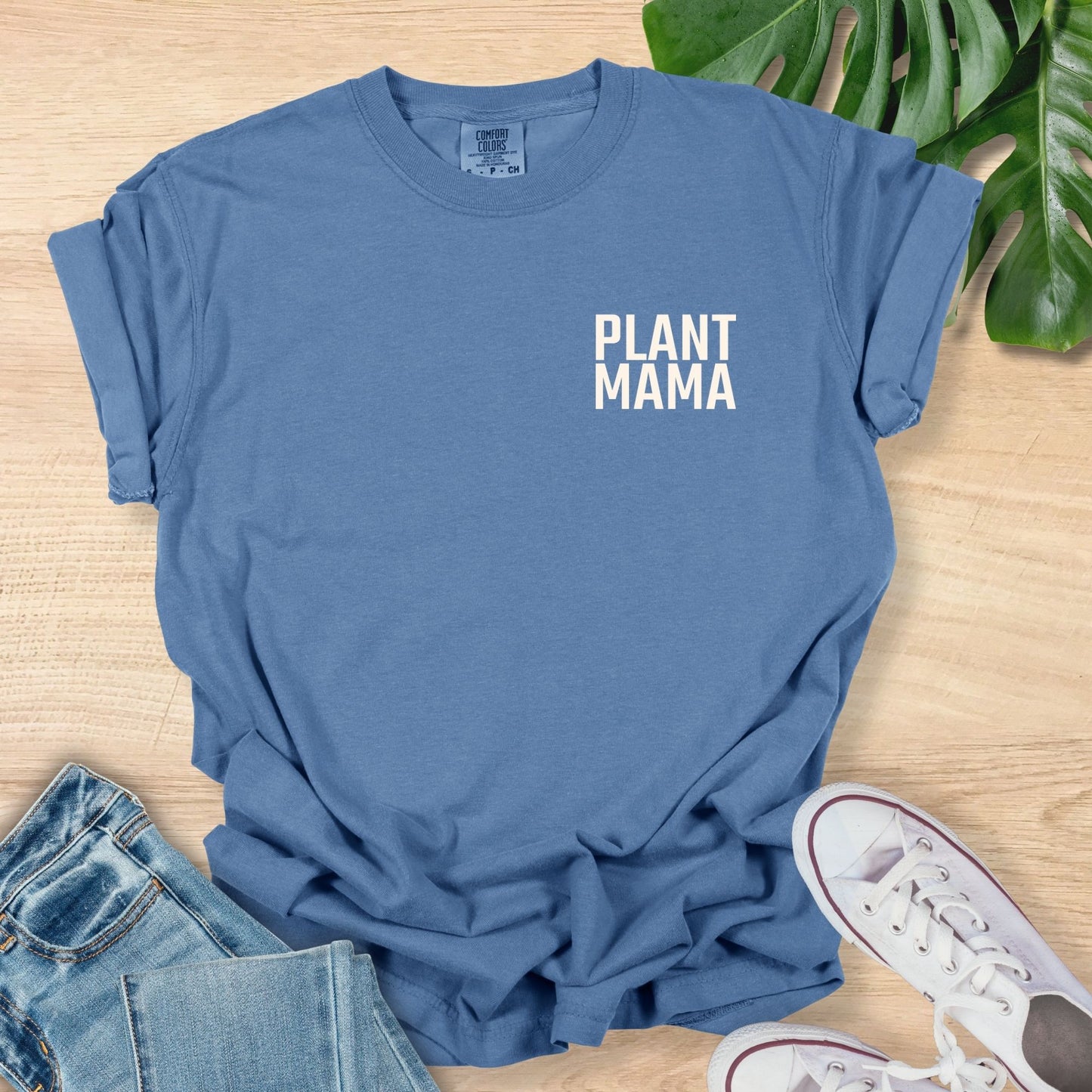 Blue t-shirt with 'PLANT MAMA' text on a wooden surface with jeans and sneakers.