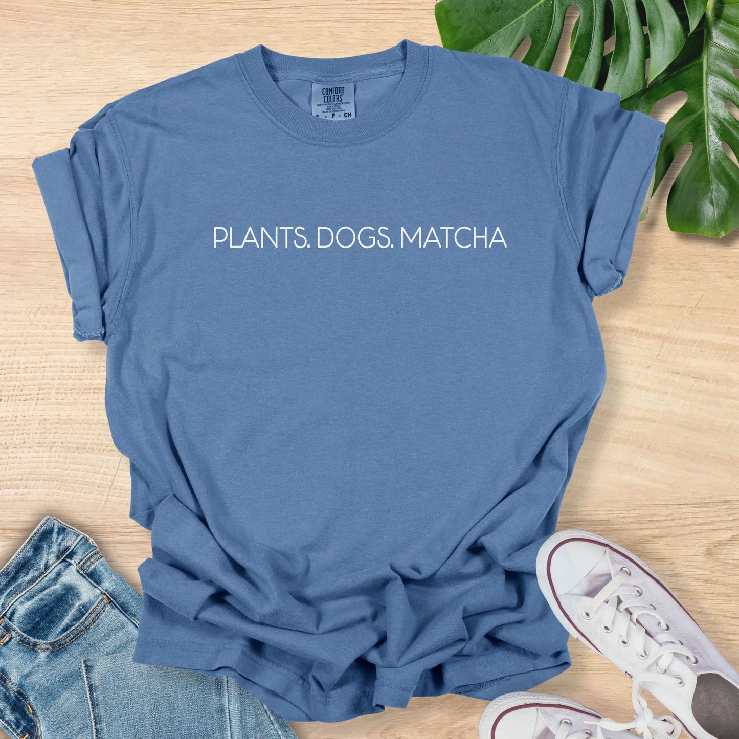 Blue t-shirt with 'PLANTS. DOGS. MATCHA' text on a wooden surface with jeans and sneakers.