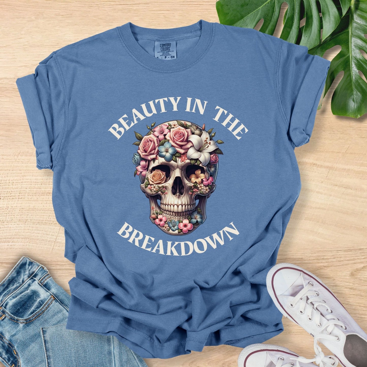 Blue t-shirt with floral skull design and text on a wooden surface with jeans and sneakers.