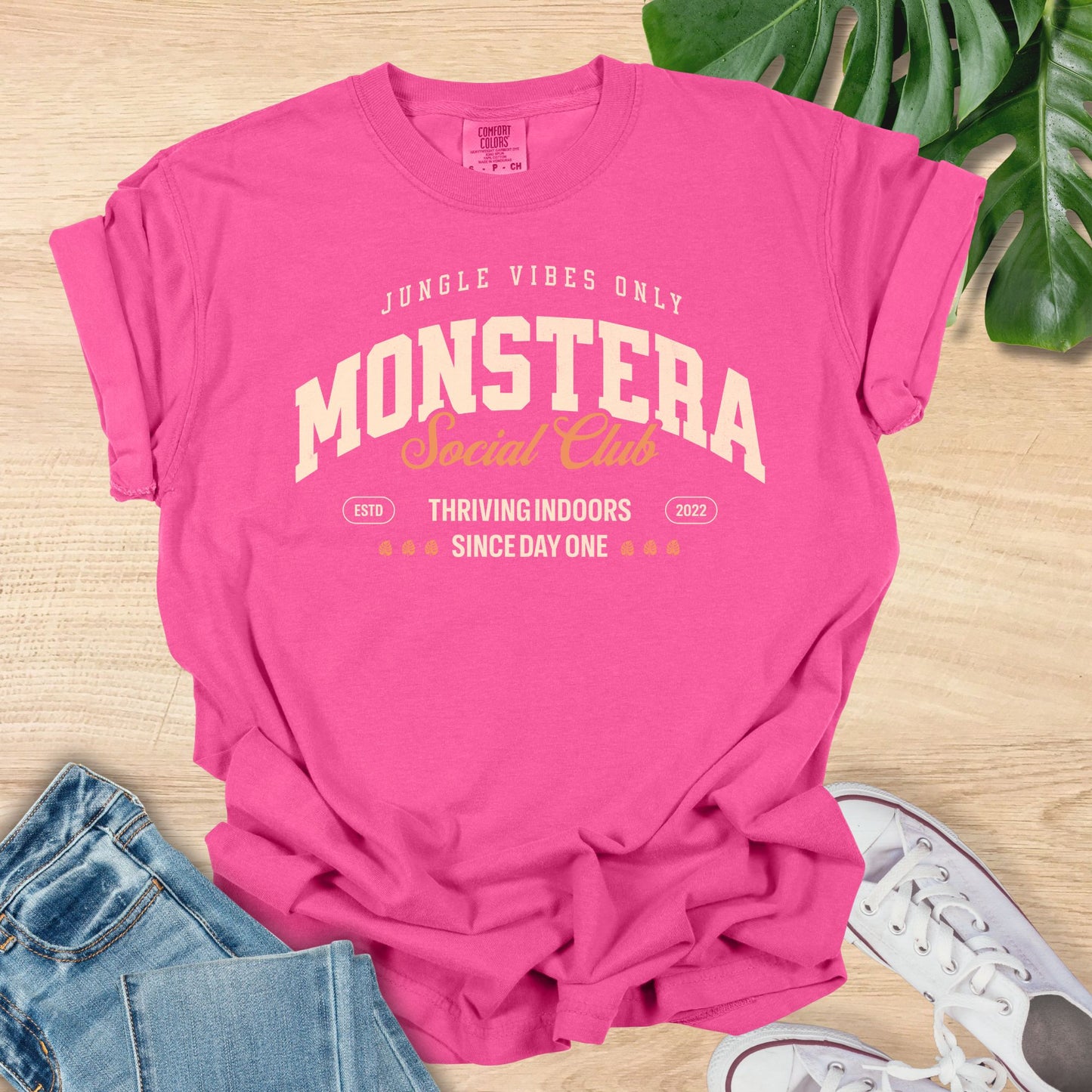 Pink t-shirt with 'Monstera Social Club' text on a wooden surface with jeans and sneakers.