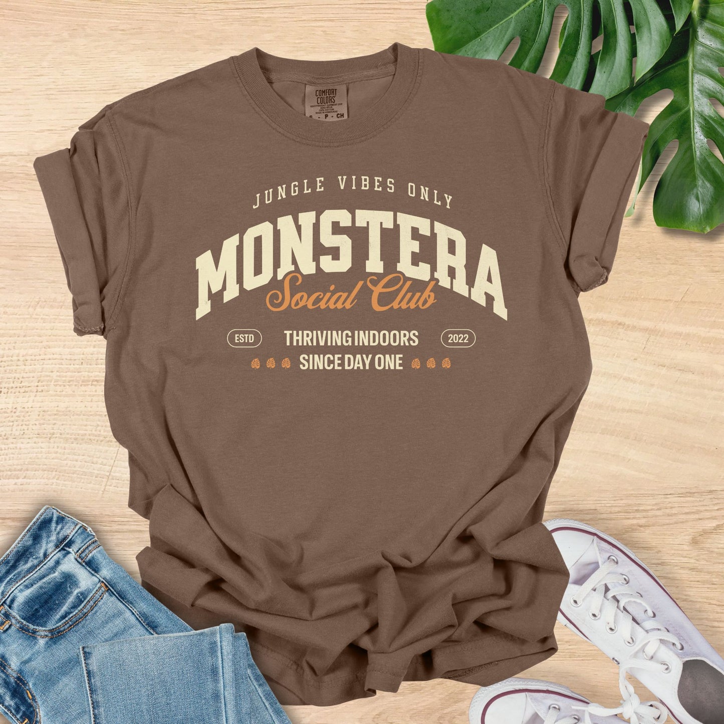 Brown t-shirt with 'Monstera Social Club' text on a wooden surface with jeans and sneakers.