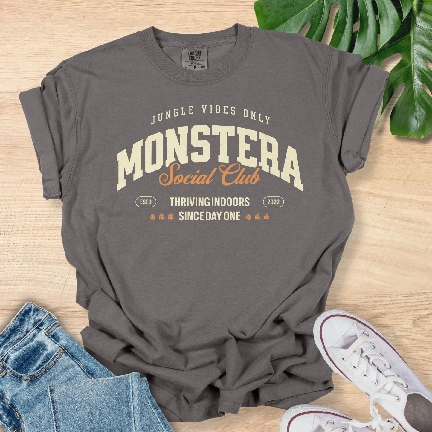 Gray t-shirt with 'Monstera Social Club' text on a wooden surface with jeans and sneakers.