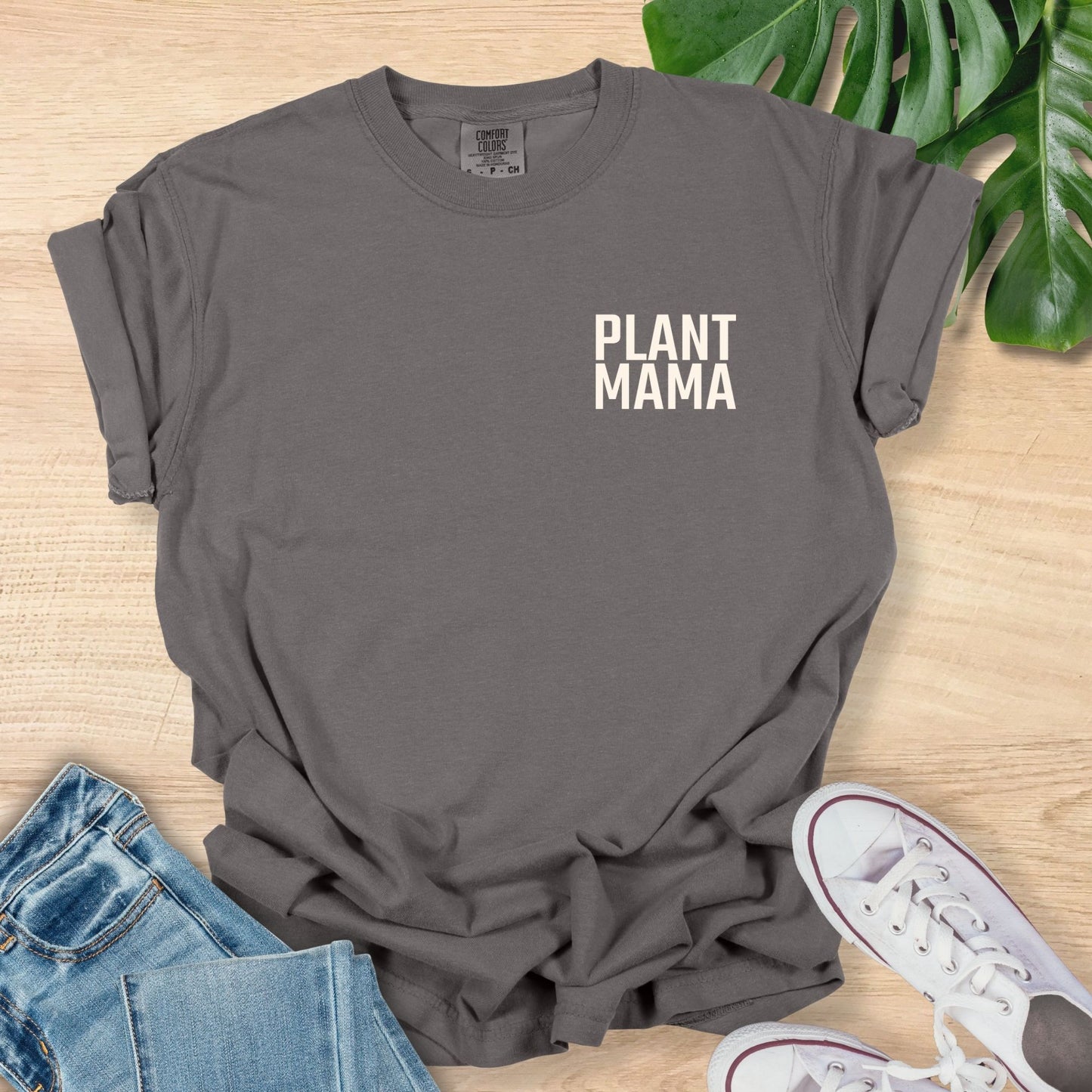 Gray t-shirt with 'PLANT MAMA' text on a wooden surface with jeans and sneakers.