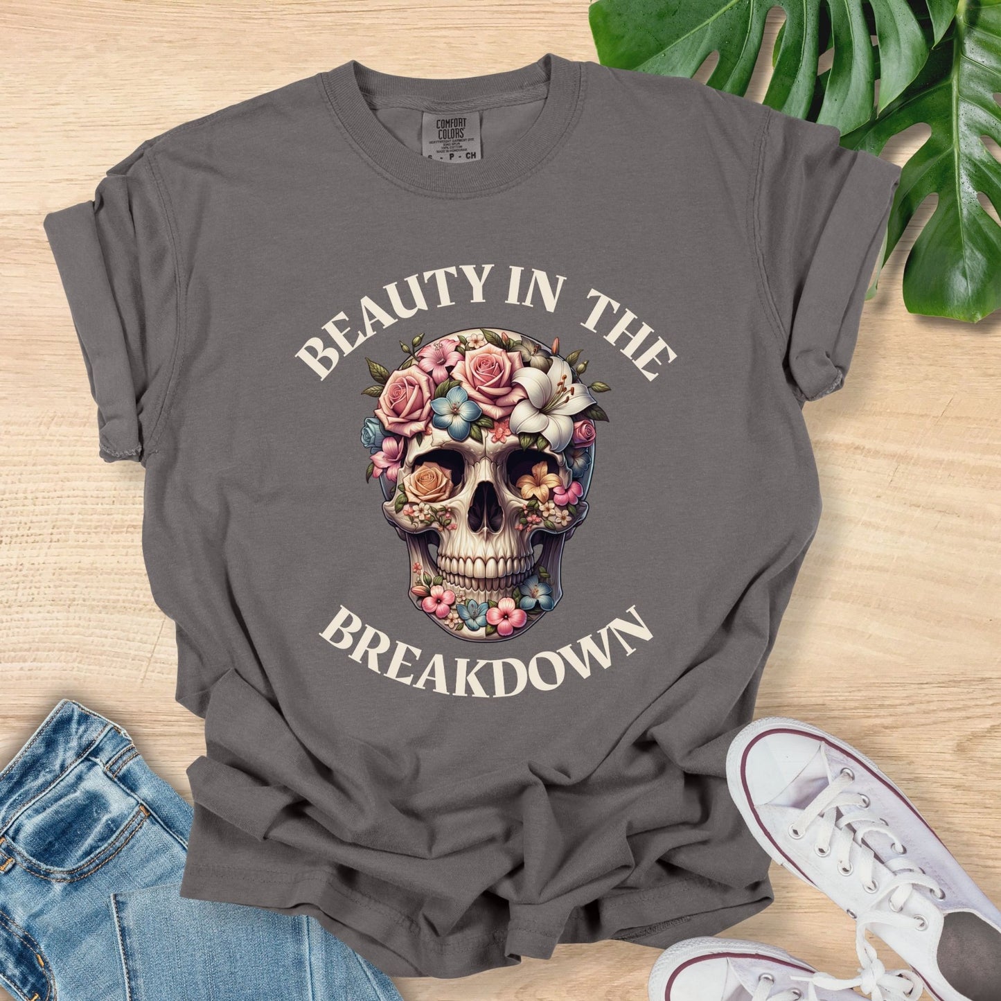 Gray t-shirt with skull and floral design on a wooden surface with jeans and sneakers.