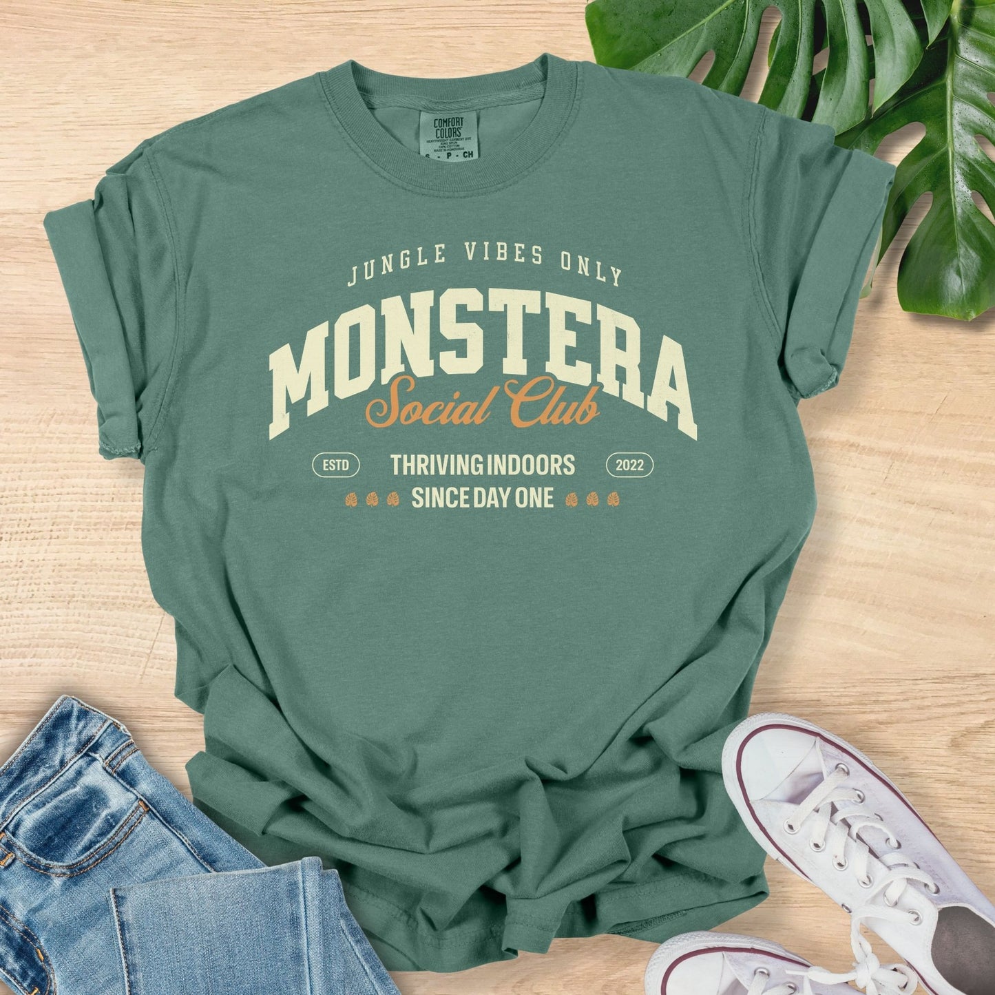 Green t-shirt with 'Monstera Social Club' text on a wooden surface with jeans and sneakers.