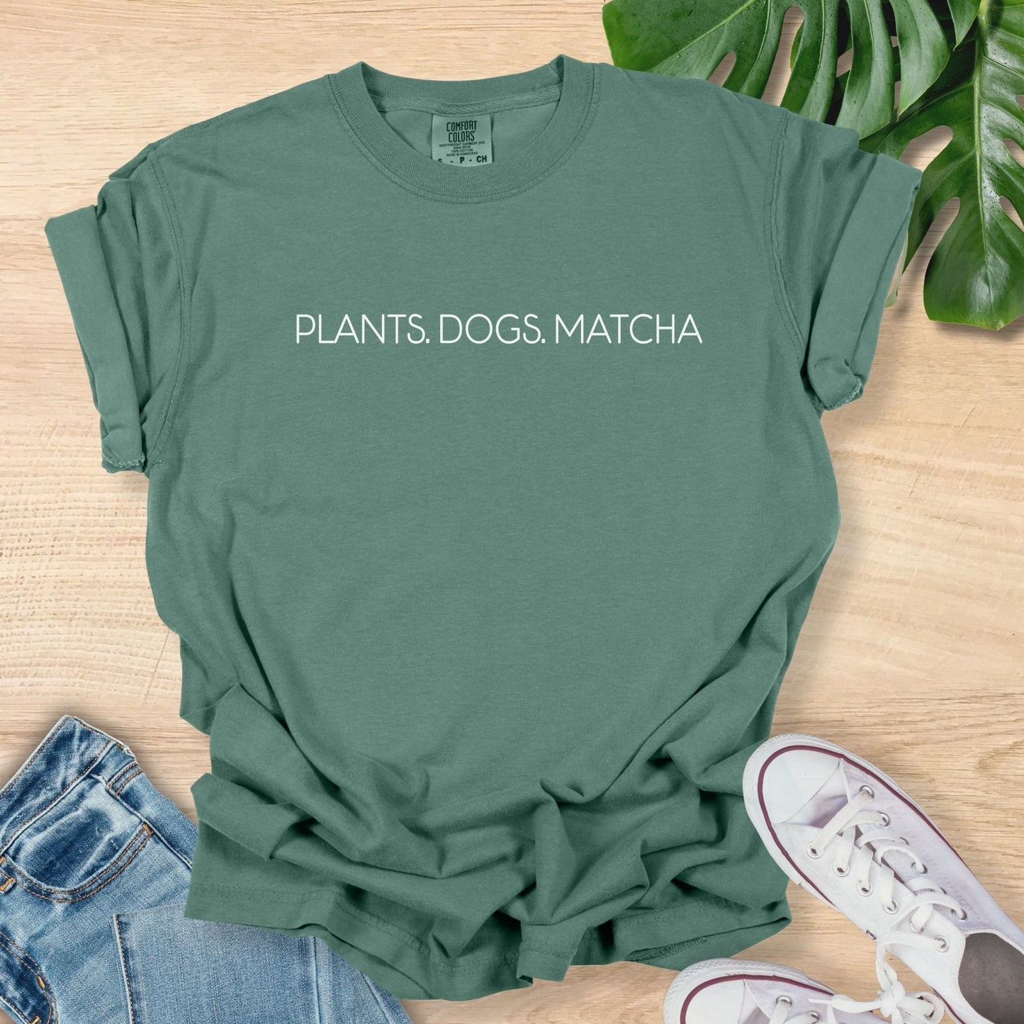 Green t-shirt with 'PLANTS. DOGS. MATCHA' text on a wooden surface with jeans and sneakers.
