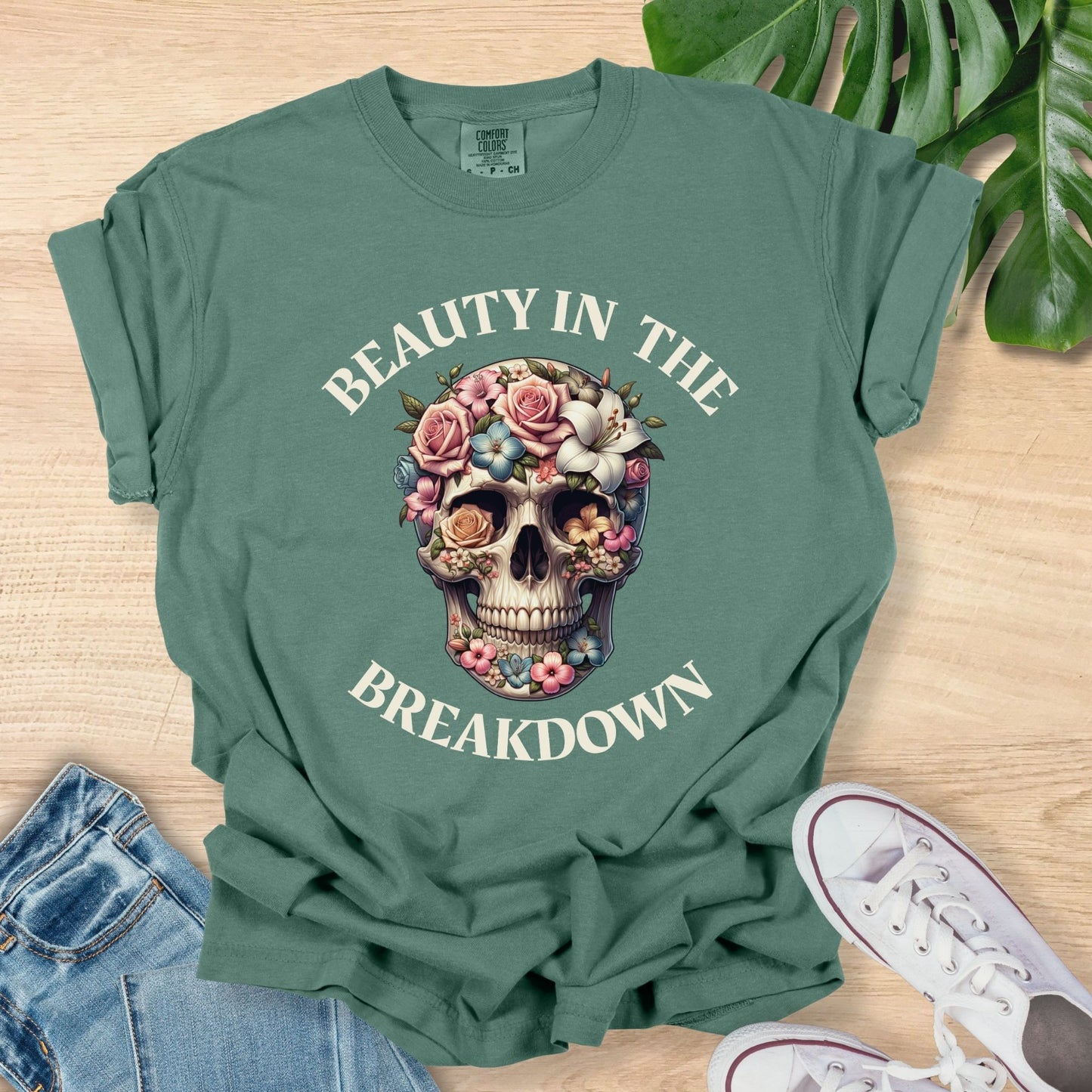 Green t-shirt with skull design and text 'Beauty in the Breakdown' on a wooden surface with jeans and sneakers.