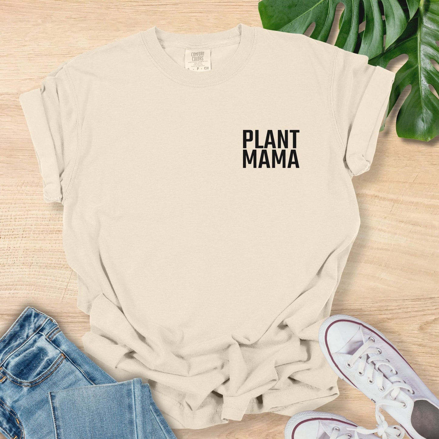 Beige t-shirt with 'PLANT MAMA' text on a wooden surface with jeans and sneakers.