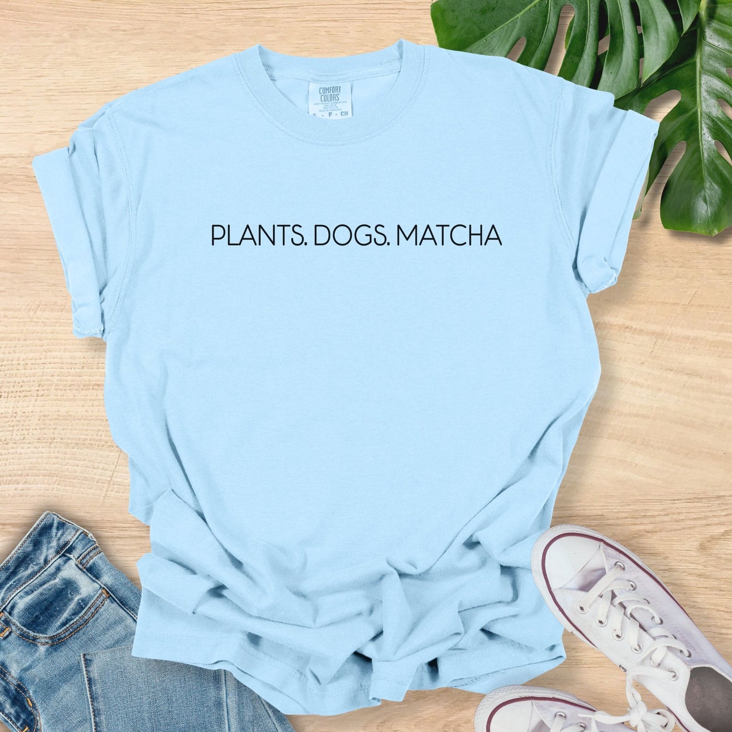 Light blue t-shirt with 'PLANTS. DOGS. MATCHA' text on a wooden surface with jeans and sneakers.