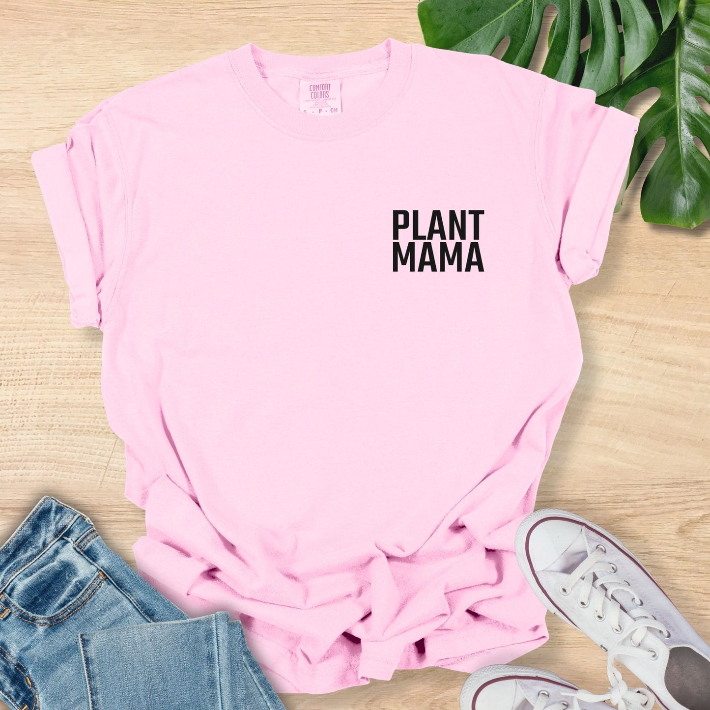 Pink t-shirt with 'PLANT MAMA' text on a wooden surface with jeans and sneakers.