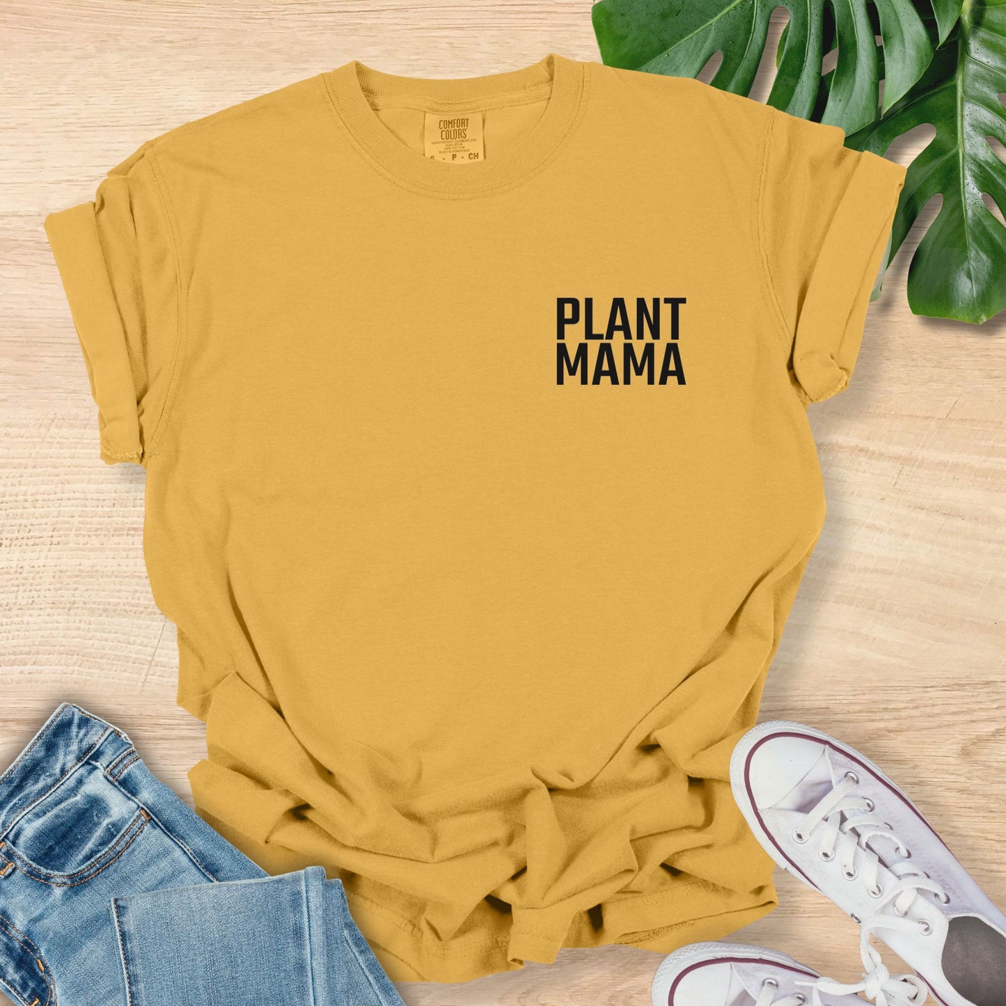 Yellow t-shirt with 'PLANT MAMA' text on a wooden surface with jeans and sneakers.