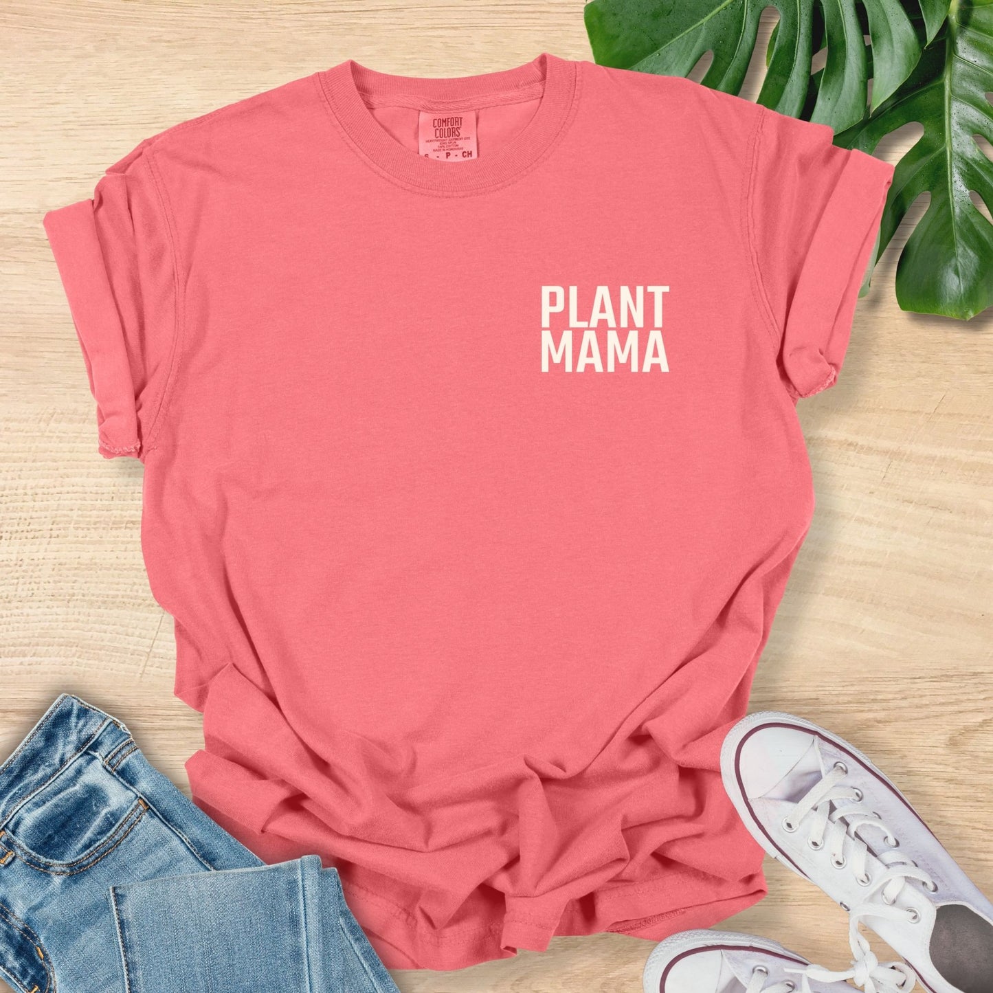 Pink t-shirt with 'PLANT MAMA' text on a wooden surface with jeans and sneakers.