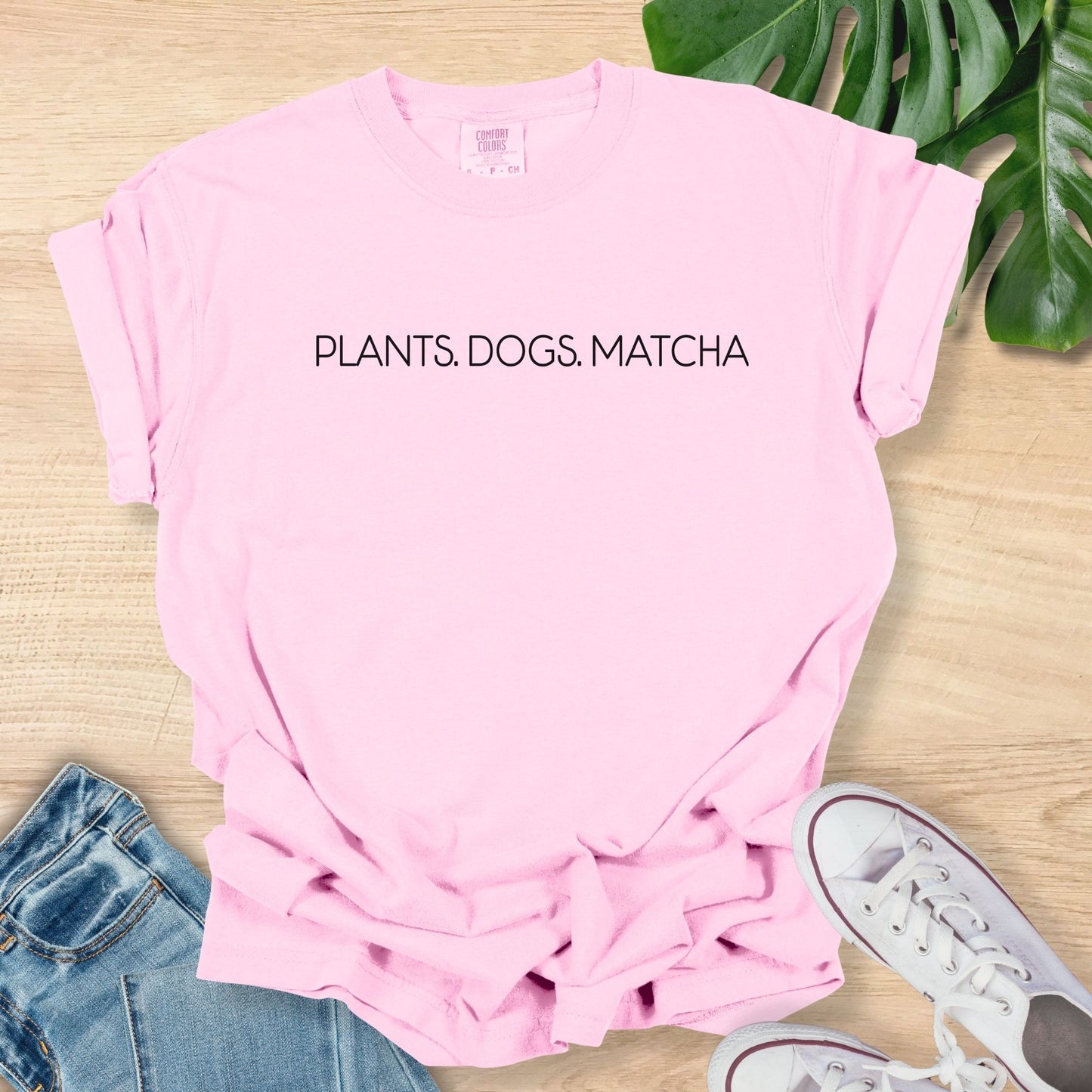 Pink t-shirt with 'PLANTS. DOGS. MATCHA' text on a wooden surface with jeans and sneakers.