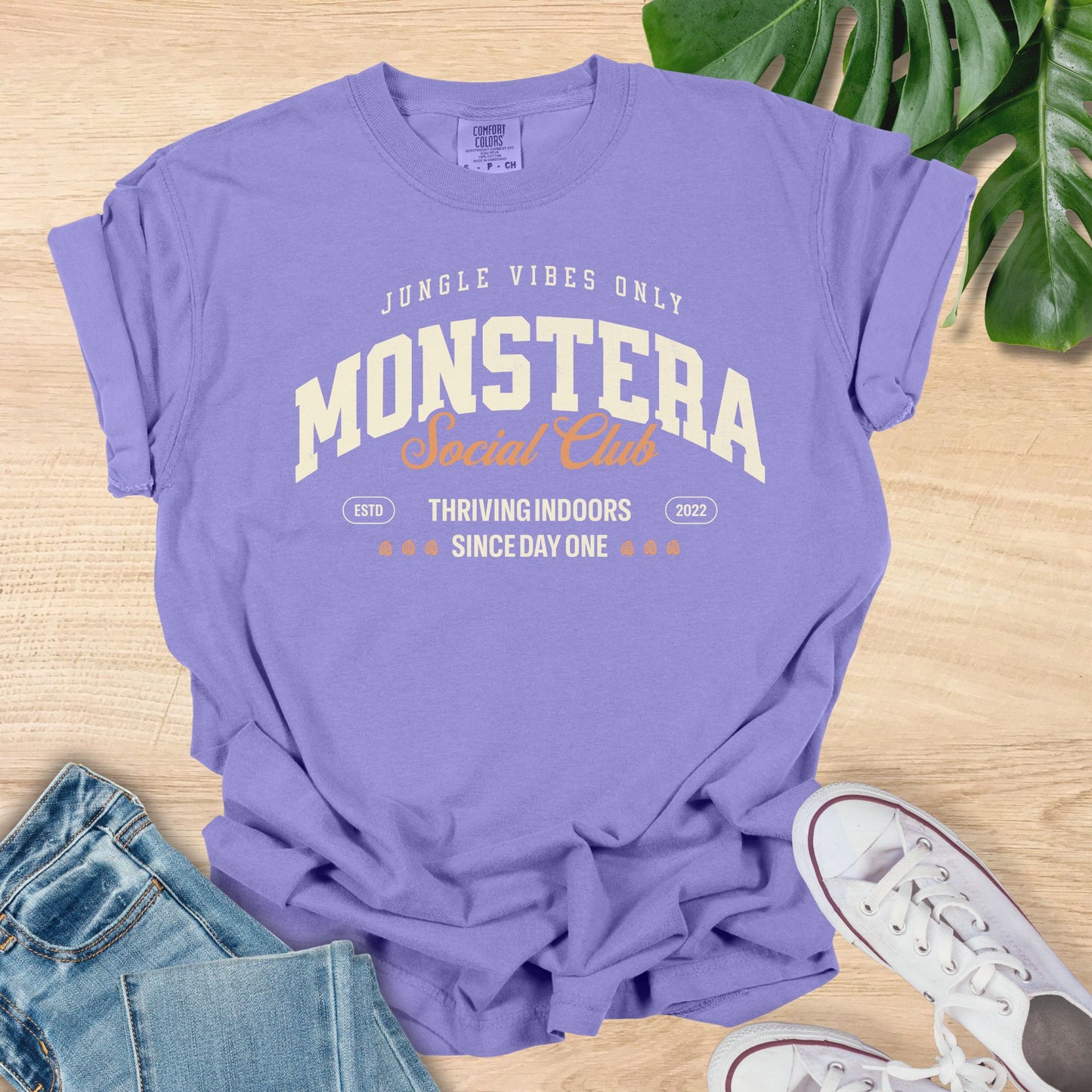 Purple t-shirt with 'Monstera Social Club' text on a wooden surface with jeans and sneakers.