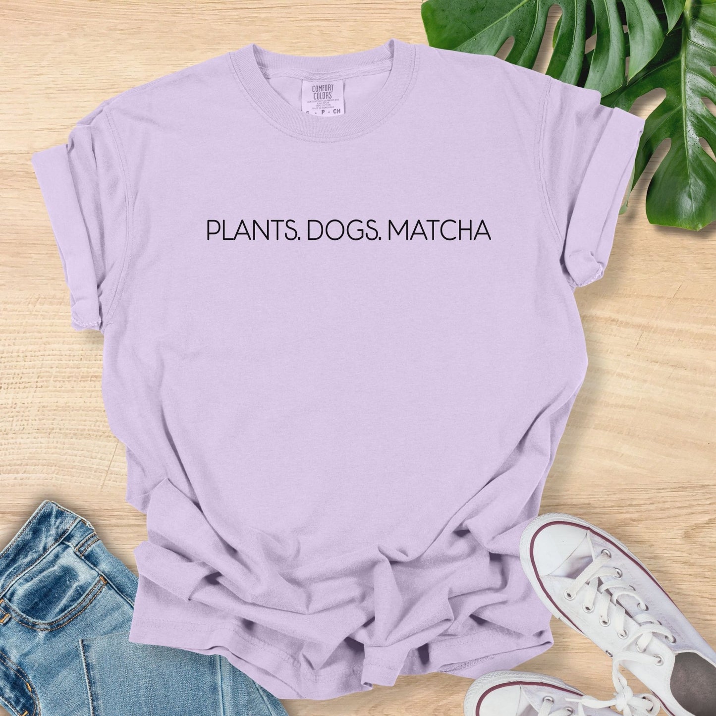 Purple t-shirt with 'PLANTS. DOGS. MATCHA' text on a wooden surface with jeans and sneakers.