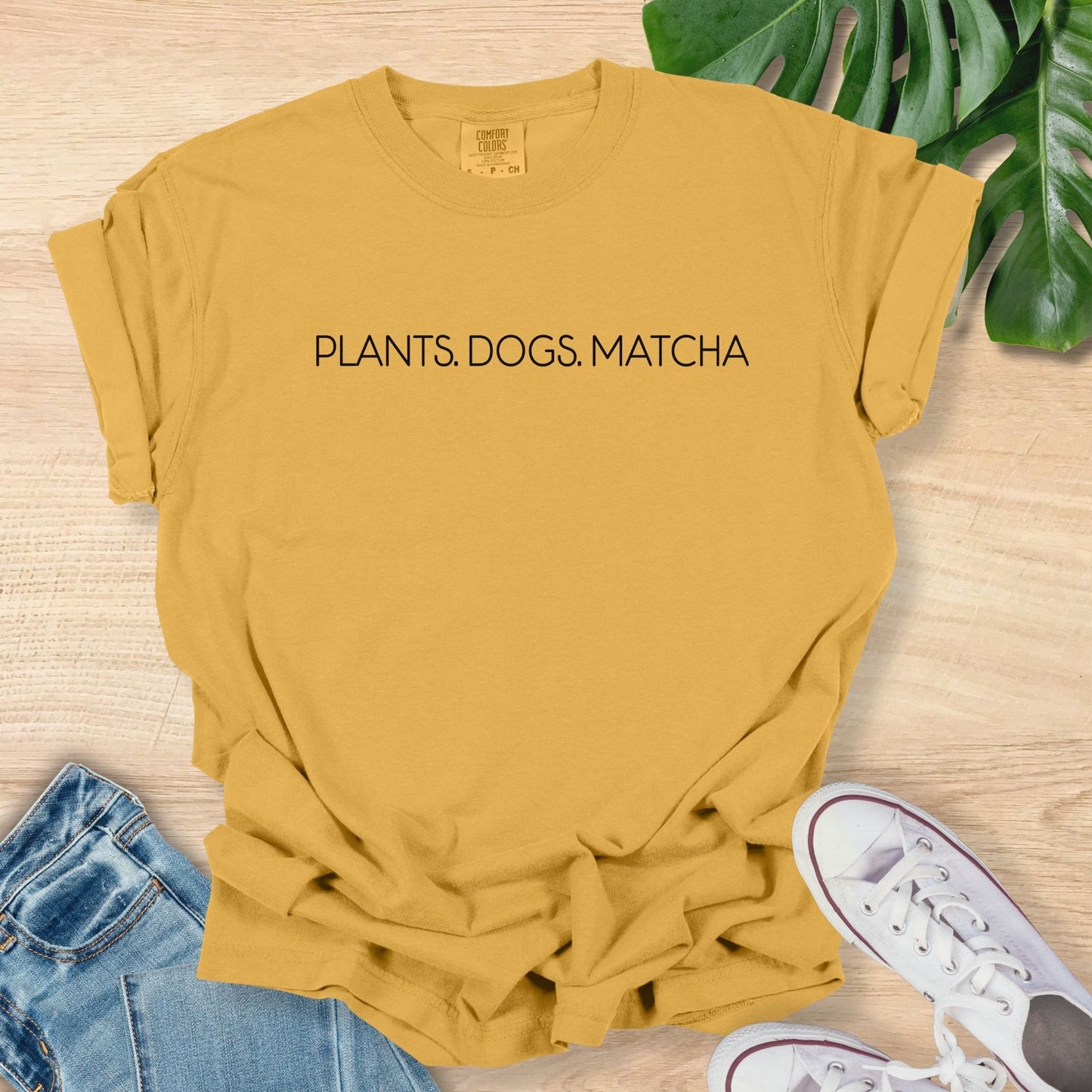 Yellow t-shirt with 'PLANTS. DOGS. MATCHA' text on a wooden surface with jeans and sneakers.