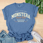 Blue t-shirt with 'Monstera Social Club' text on a wooden surface with jeans and sneakers.