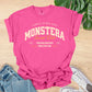 Pink t-shirt with 'Monstera Social Club' text on a wooden surface with jeans and sneakers.