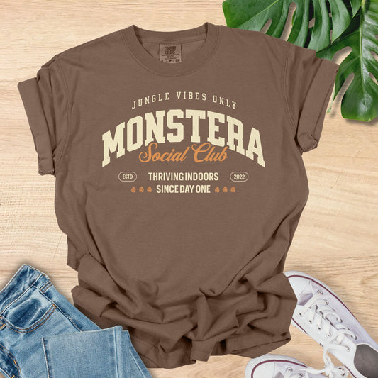 Brown t-shirt with 'Monstera Social Club' text on a wooden surface with jeans and sneakers.