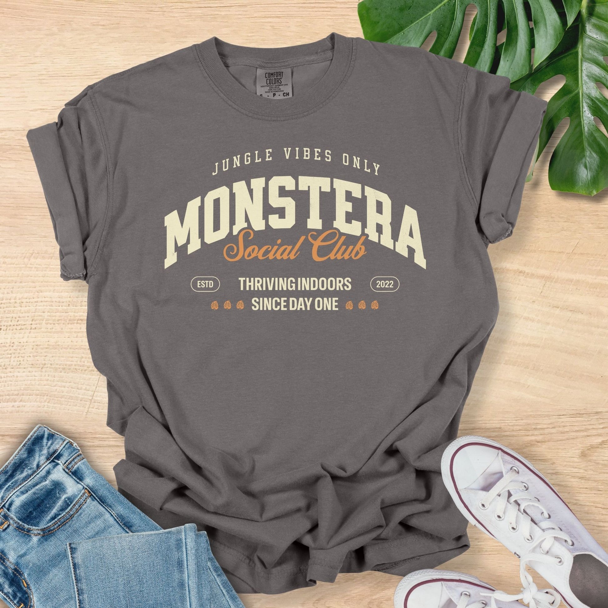 Gray t-shirt with 'Monstera Social Club' text on a wooden surface with jeans and sneakers.