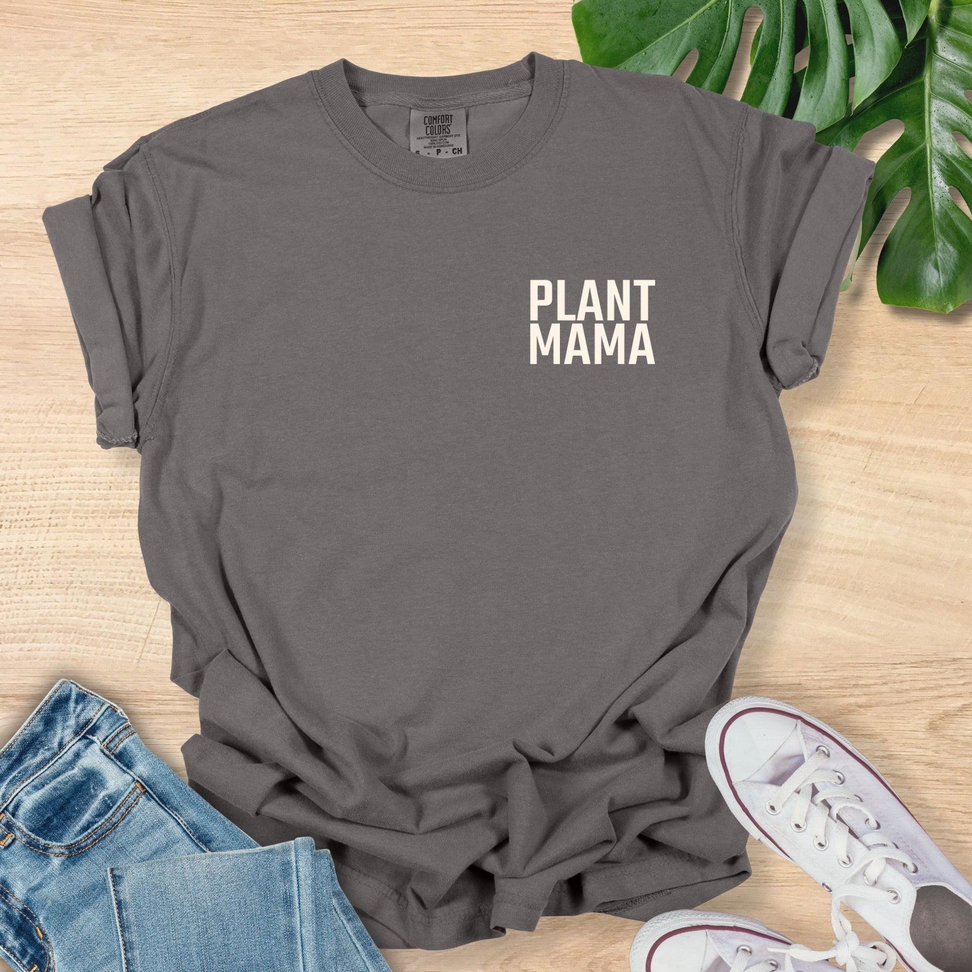 Gray t-shirt with 'PLANT MAMA' text on a wooden surface with jeans and sneakers.
