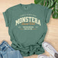 Green t-shirt with 'Monstera Social Club' text on a wooden surface with jeans and sneakers.