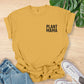 Yellow t-shirt with 'PLANT MAMA' text on a wooden surface with jeans and sneakers.