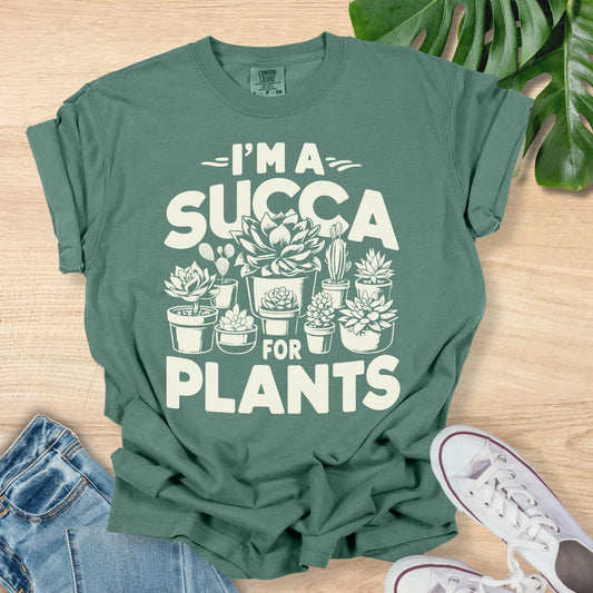 Green t-shirt with 'I'm a Succa for Plants' design on a wooden surface with plants and jeans.